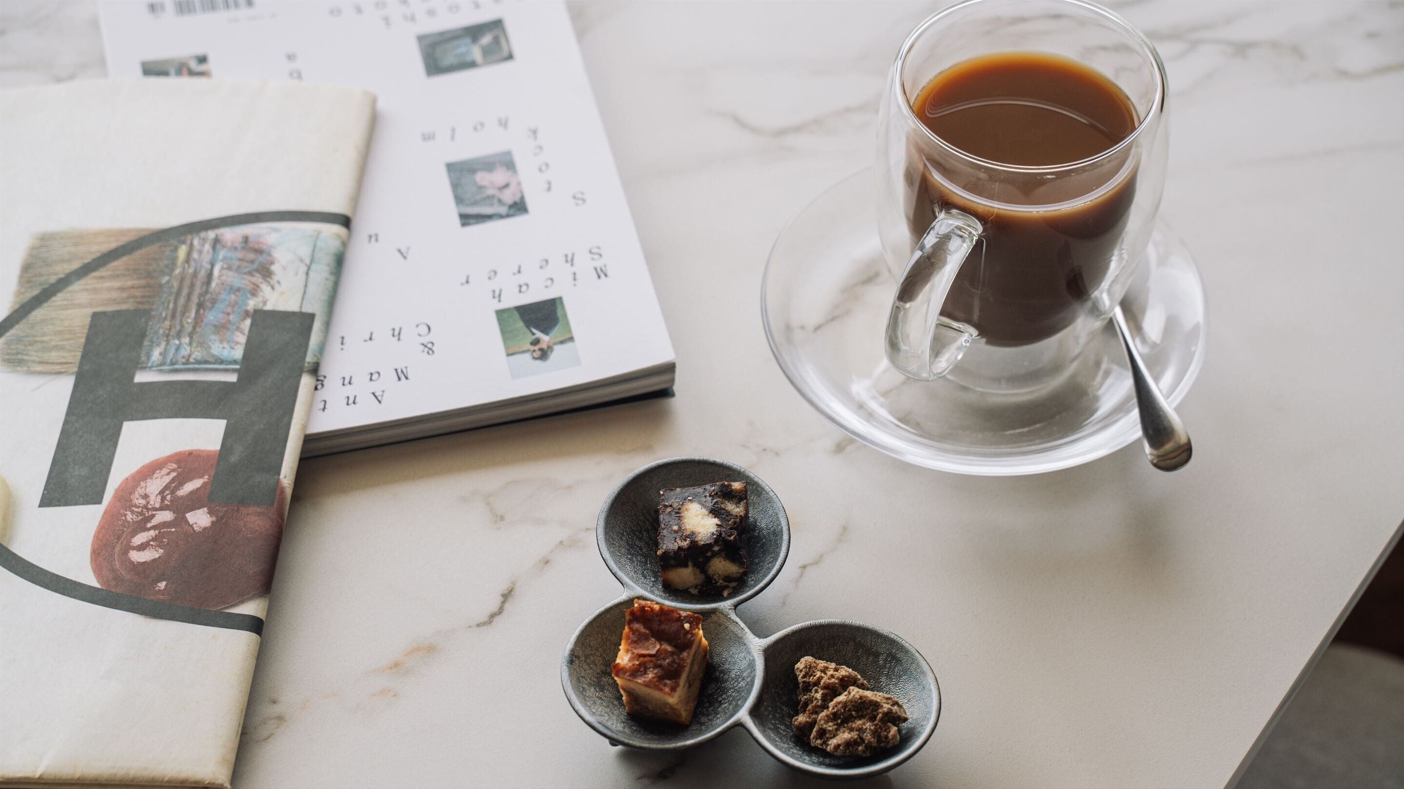 A clear glass cup of coffee on a saucer, a magazine with visible text and images, and three small dishes with food items on a marble surface.