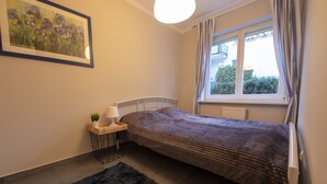 A bedroom with a bed, a bedside table with a lamp, a small plant, a window with curtains, and a painting on the wall. - Victus Apartamenty - Avocado (Sopot)