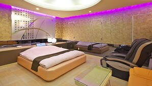 Une chambre d’hôtel moderne avec un grand lit, un canapé, un fauteuil massant et une table en verre.