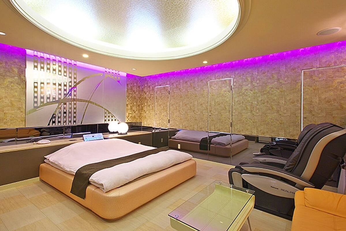 A modern hotel room with a large bed, a sofa, a massage chair, and a glass table.
