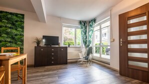 A modern room with a wooden floor, a dining table with chairs, a TV on a cabinet, and a window with curtains. - Victus Apartamenty - Bella (Sopot)