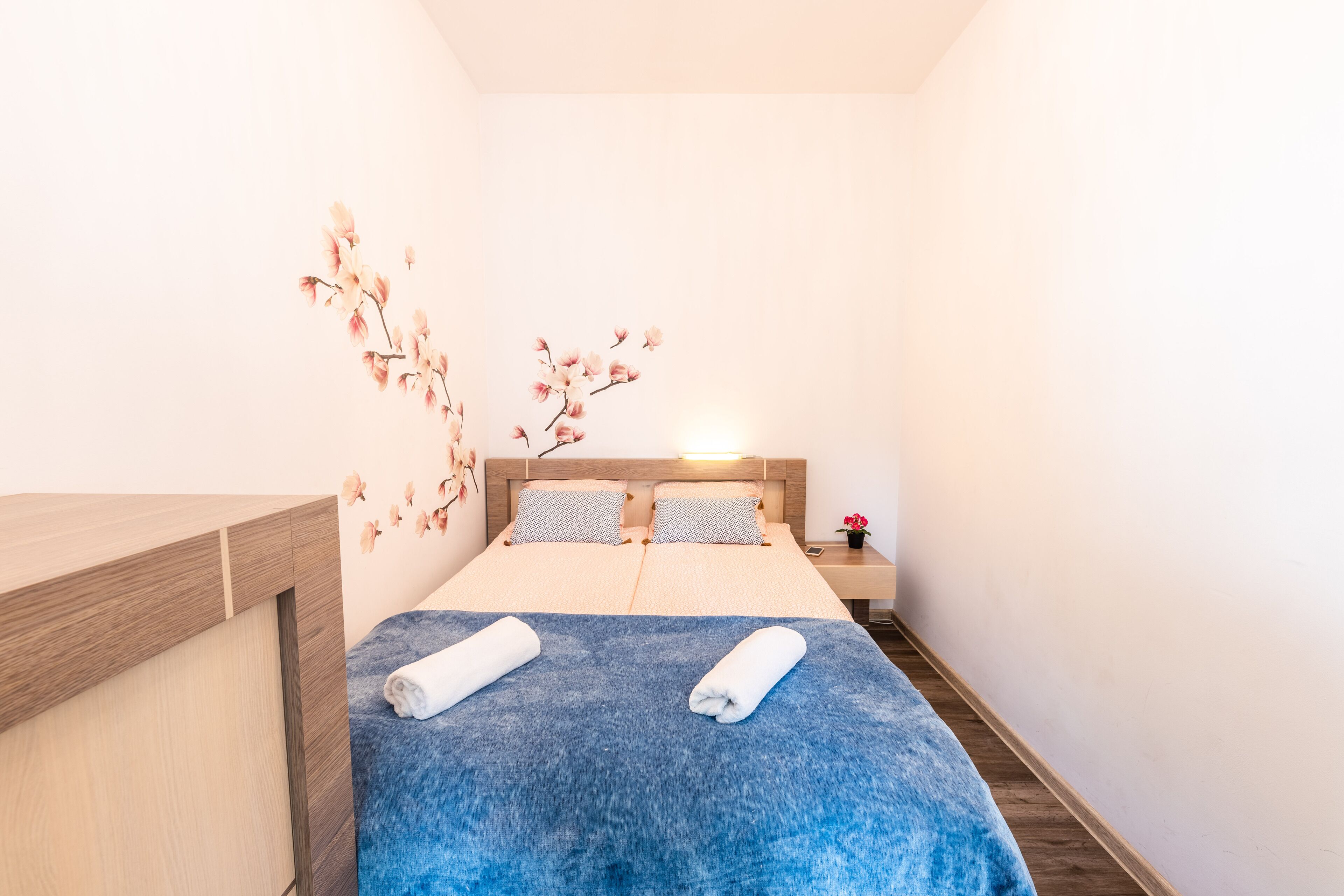 Ett hotellrum med en säng, ett nattduksbord i trä, en vägglampa och en dekorativ blommotiv på väggen.