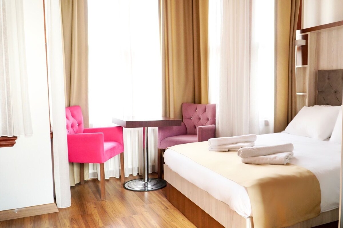 Ein Hotelzimmer mit einem Bett, einem rosa Sessel, einem gesteppten Ohrensessel und einem kleinen Tisch.