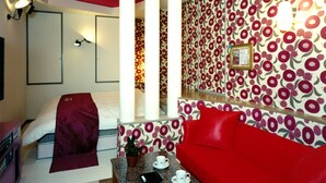 A hotel room with a red sofa, a small table with a plant and cups, a bed with a red blanket, and a wall with a floral pattern.