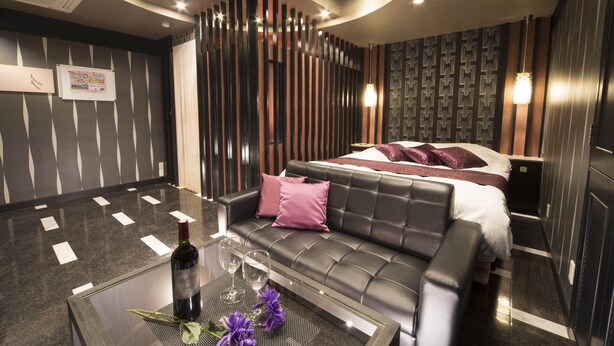 A modern hotel room with a leather sofa, a glass coffee table with a bottle and flowers, a bed with a patterned headboard, and a decorative wall panel. - Hotel Dion -dreams IN Resort - Adults Only (Miyoshi)