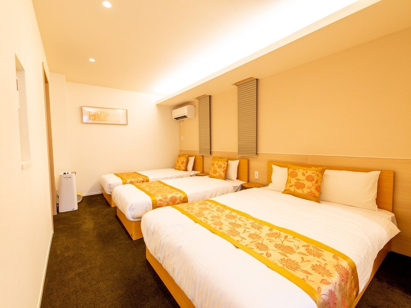 A hotel room with three beds, a small bedside table, an air conditioning unit, and a framed picture on the wall.