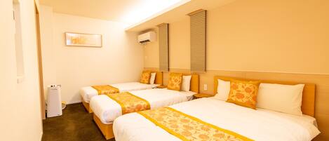 A hotel room with three beds, each with white bedding and orange floral bedspreads. There is a small bedside table with a lamp, a wall-mounted air conditioner, and a framed picture on the wall.