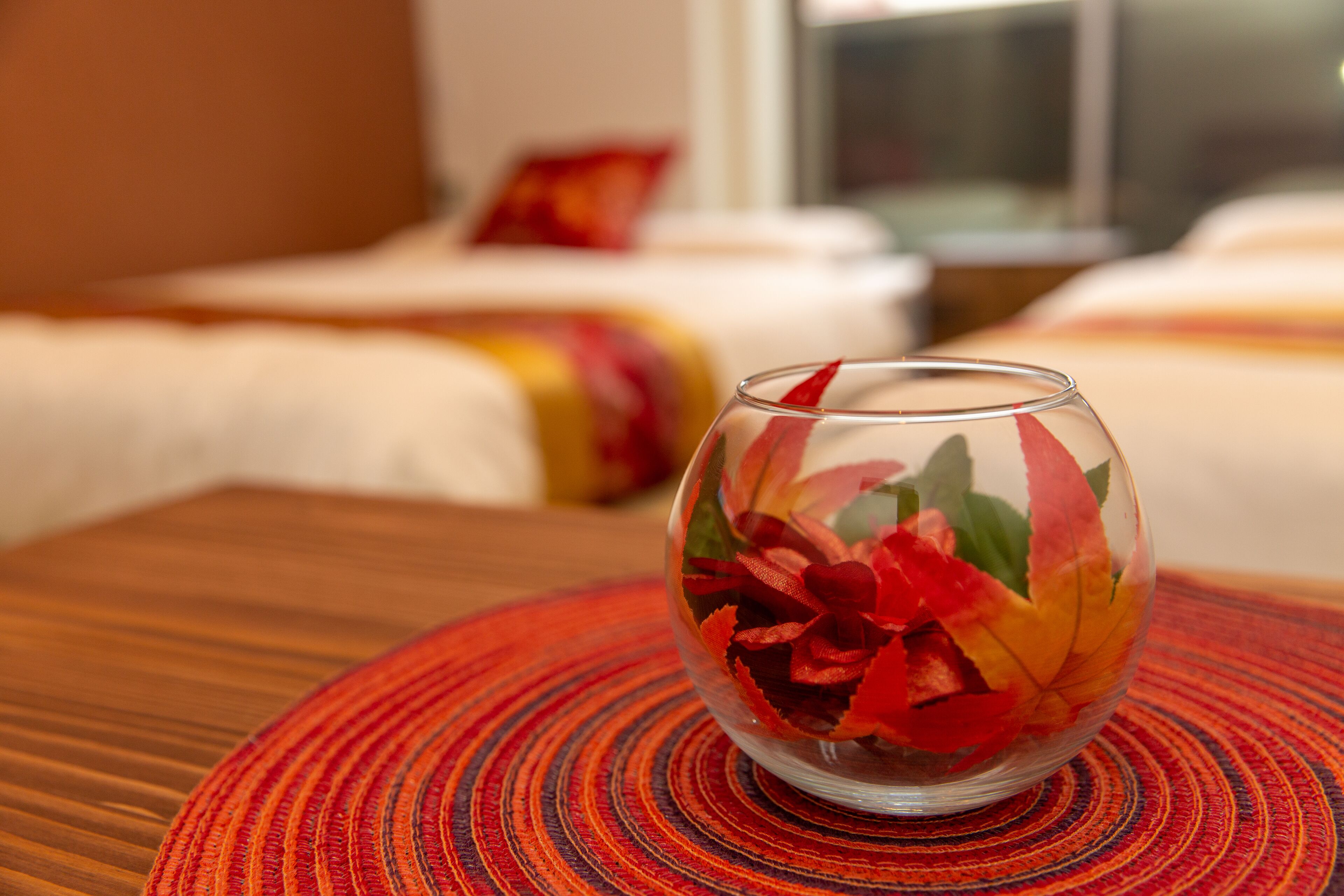A clear glass bowl with red and yellow leaves on a wooden table in a hotel room.
