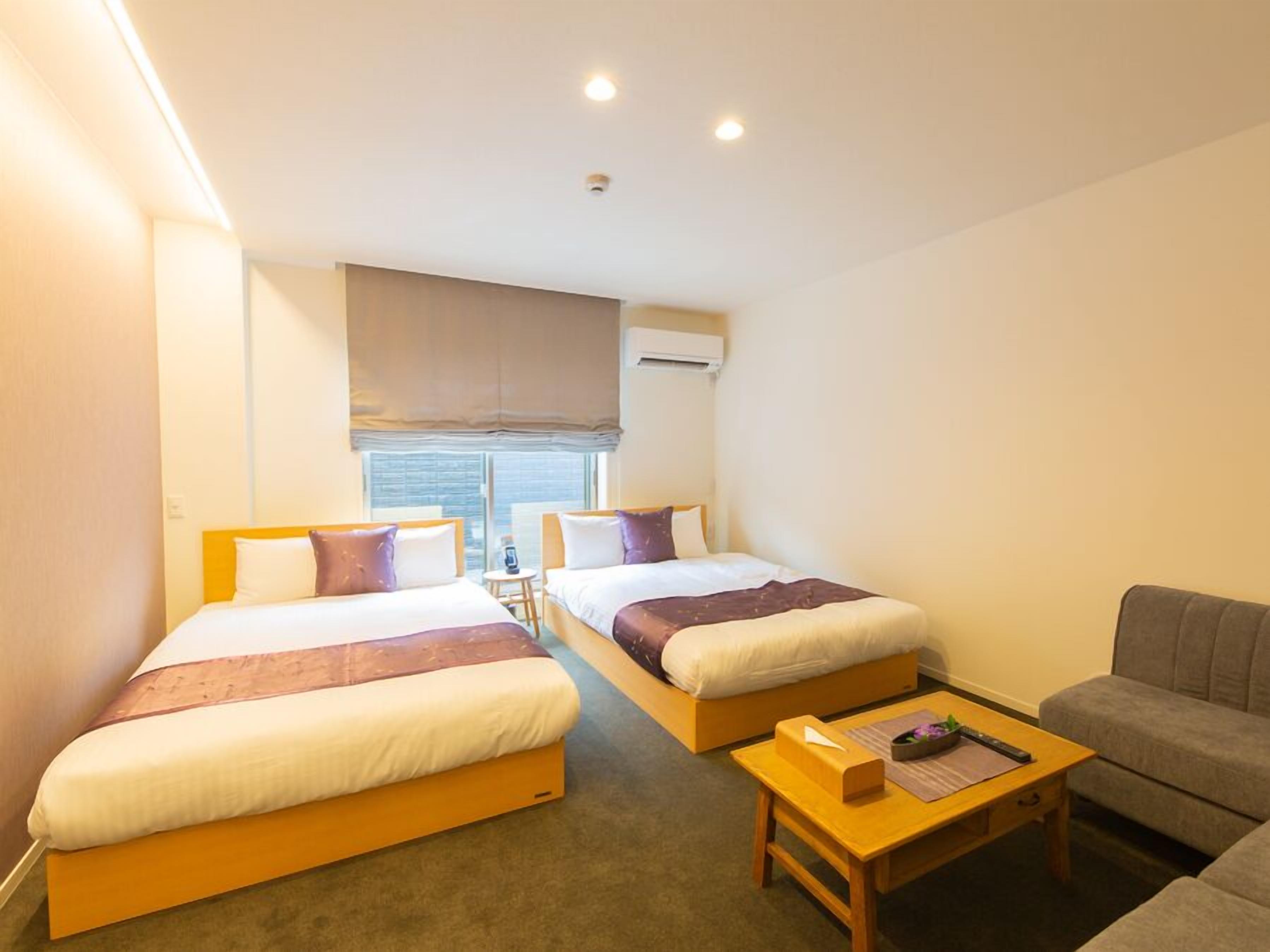 A hotel room with two beds, a small table, a sofa, and a window with blinds.