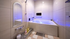 Une salle de bain moderne dotée d’une grande baignoire, d’une douchette à main, d’un meuble-lavabo avec un miroir et d’un téléviseur fixé au mur.