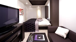 Une chambre d’hôtel moderne dotée d’un grand téléviseur à écran plat, d’un lit avec une couvre-lit à motifs, d’un canapé avec un coussin blanc et d’une petite table sur laquelle se trouvent un vase et un livre.