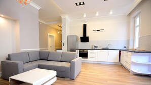 Una sala moderna con un sofá seccional gris y una mesa de centro blanca. Se ve una cocina con gabinetes blancos y una campana extractora negra al fondo.