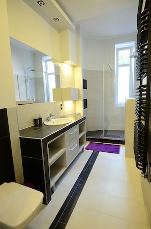 A modern bathroom with a white sink, mirror, and toilet. There is a shower area with a glass door and a window providing natural light. - Victus Apartamenty - Belweder (Sopot)