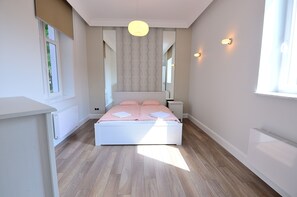A bedroom with a white bed, wooden flooring, a large window, and a ceiling light. - Victus Apartamenty - Belweder (Sopot)