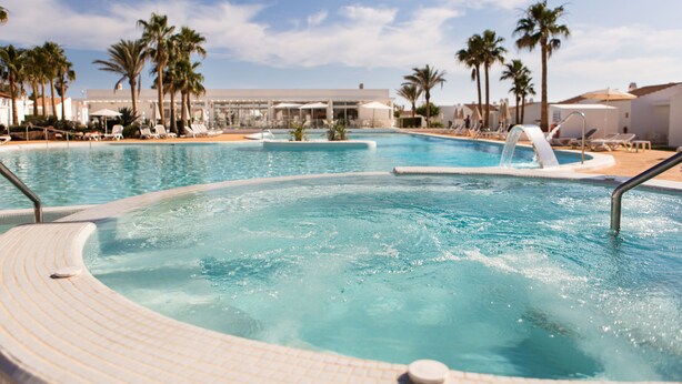 Una amplia piscina al aire libre con varias pistas, rodeada de palmeras y sillas de descanso.