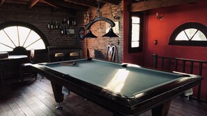 Une table de billard dans une pièce au plancher en bois et aux murs en briques, avec un porte-billes et un luminaire suspendu.