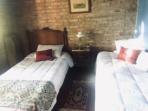 A room with two beds, a brick wall, a wooden headboard, a nightstand with a lamp, and a patterned rug.