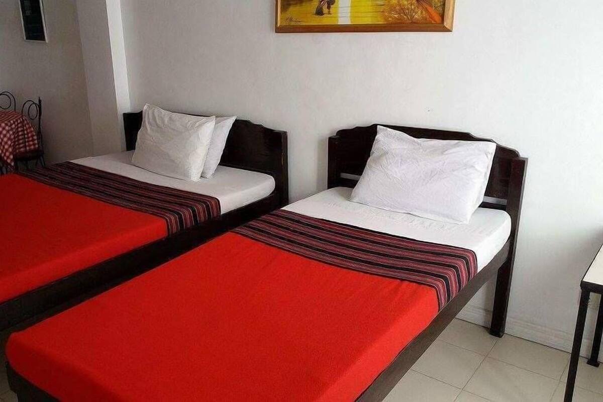 Deux lits simples avec une literie à rayures rouges et noires, une petite table et une chaise recouverte d’une nappe à carreaux.