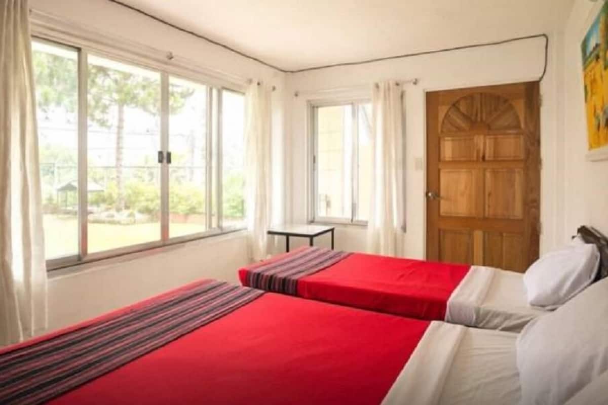 Une chambre avec deux lits, de grandes fenêtres, une petite table et une porte en bois.