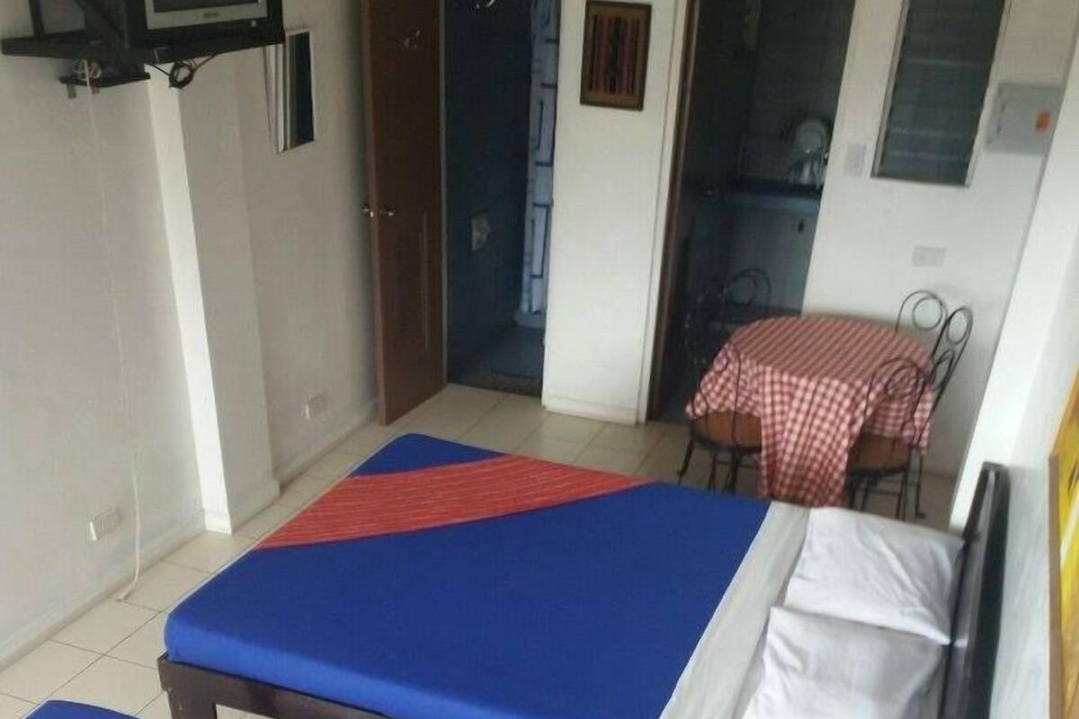 Deux lits simples avec une literie bleue et rouge, une petite table recouverte d’une nappe à carreaux et un téléviseur fixé au mur.