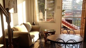 A living room with a sofa, a dining table with chairs, a staircase, and a view of buildings through a window. - Nardi-Mar Transient House (Baguio)
