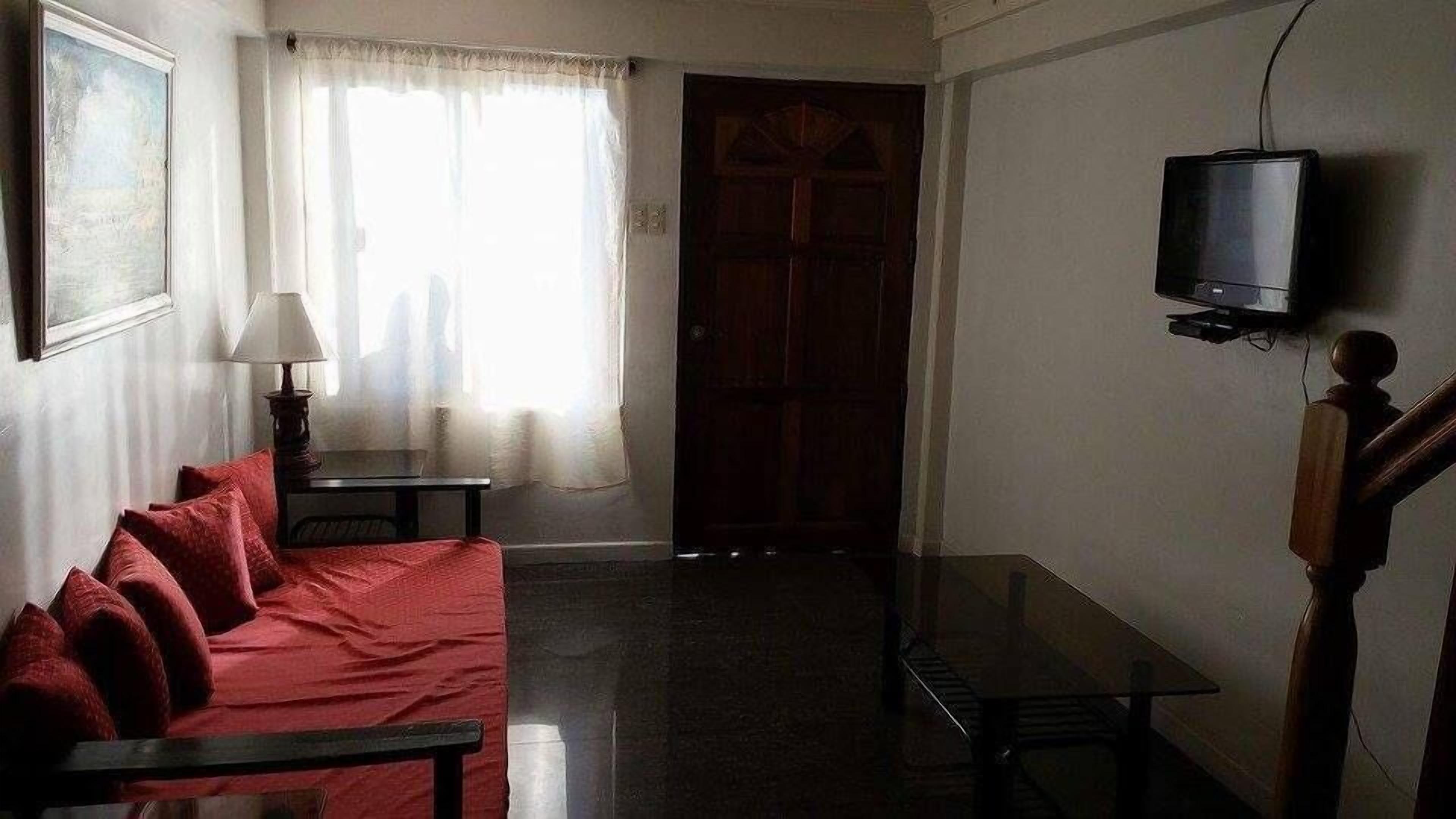 Une pièce avec un canapé rouge, une petite table, une lampe, une porte en bois et un téléviseur fixé au mur.