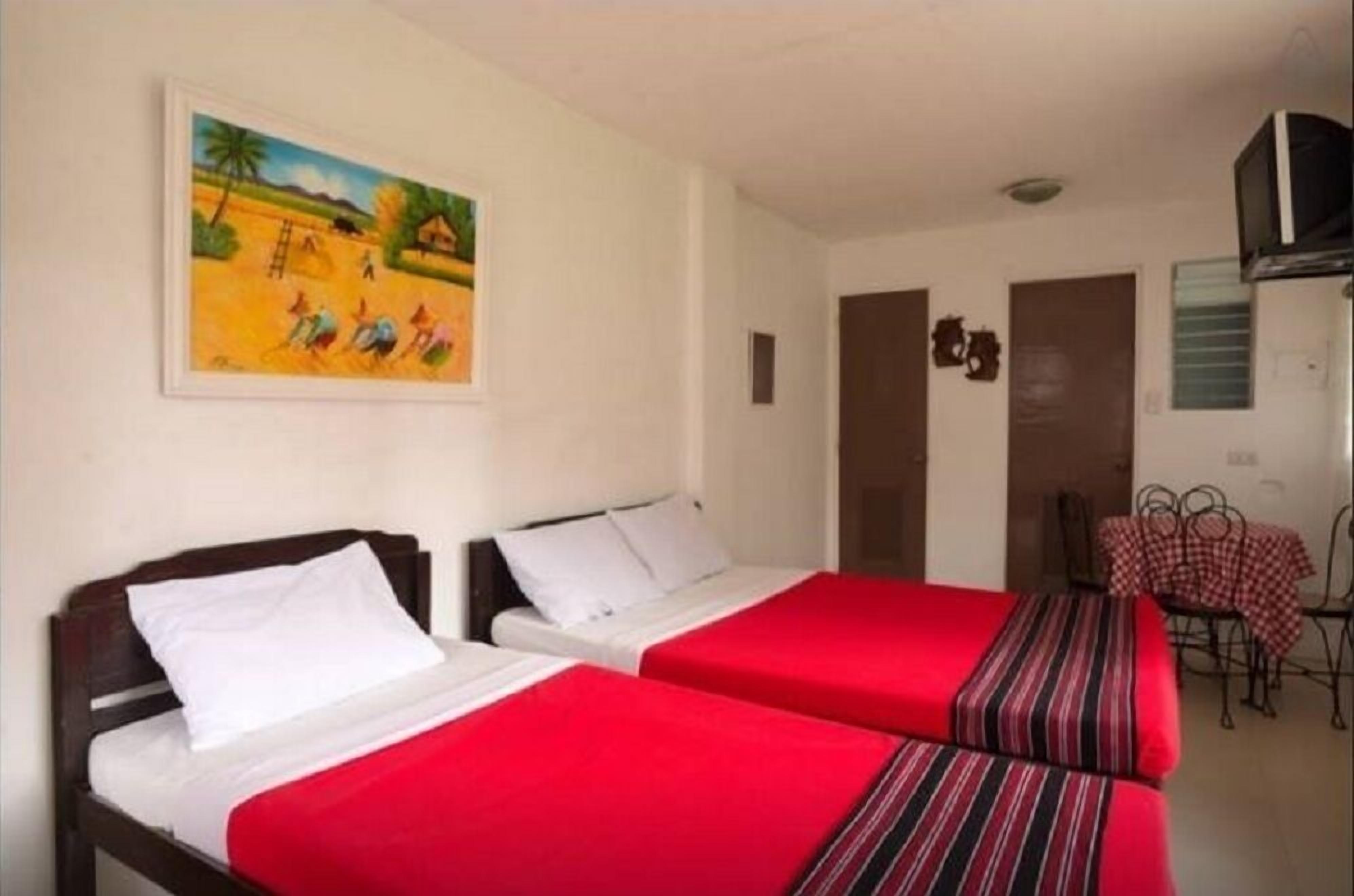 Ein Zimmer mit zwei Betten, jeweils mit roter Bettdecke und weißen Kissen. An der Wand hängt ein Bild, das eine ländliche Szene zeigt. In der Ecke steht ein kleiner Tisch mit kariertem Tuch.