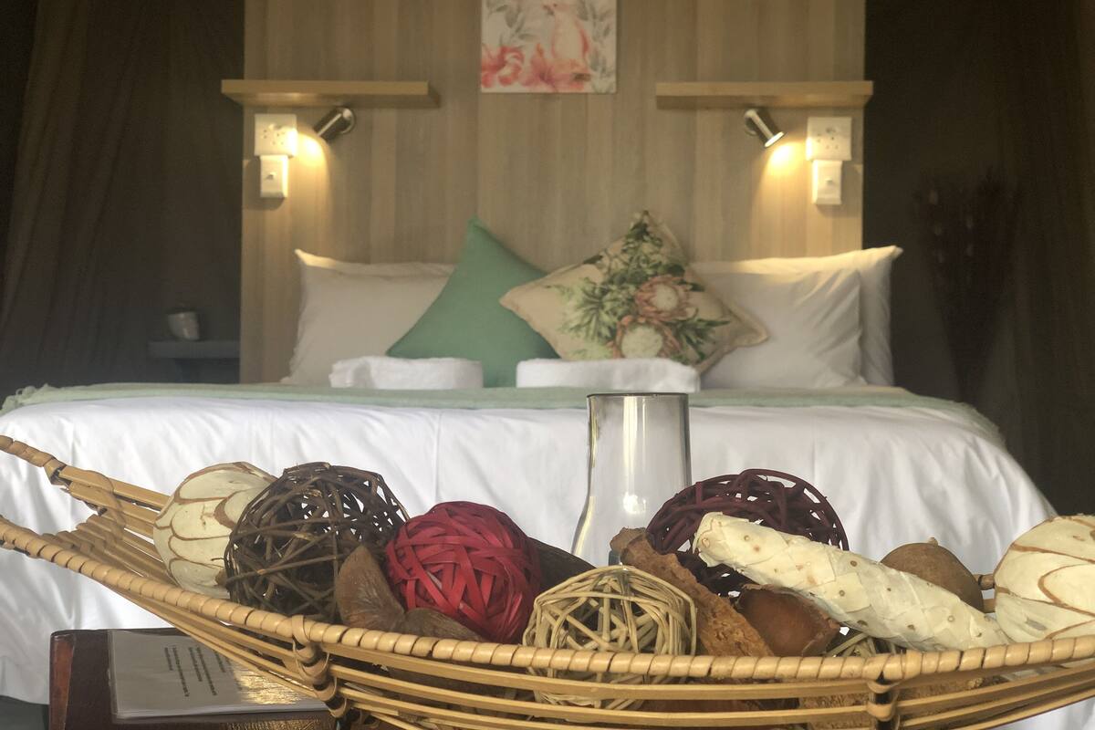 A neatly made bed with white linens, a green pillow, and a decorative pillow with a floral design. There is a wicker basket filled with various fruits and vegetables on the bed.