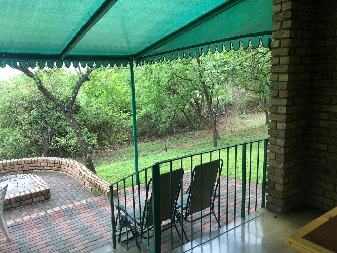 A covered outdoor seating area with brick flooring and metal railings, overlooking a grassy landscape with trees.