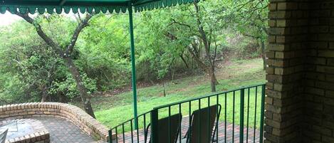 A covered outdoor seating area with brick flooring and metal railings, overlooking a grassy landscape with trees.