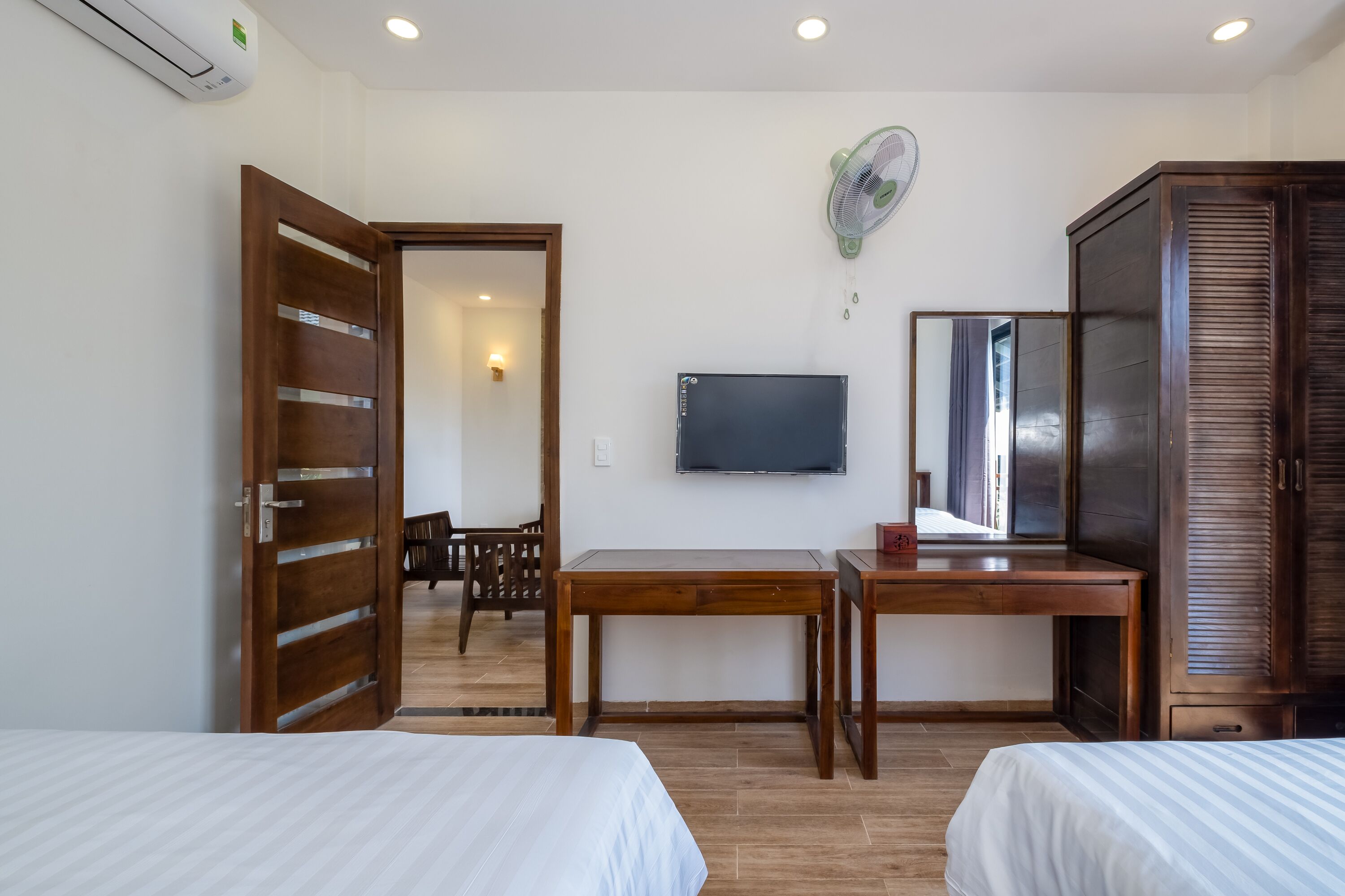 A hotel room with two beds, a wooden desk, a television, a wardrobe, and a fan.