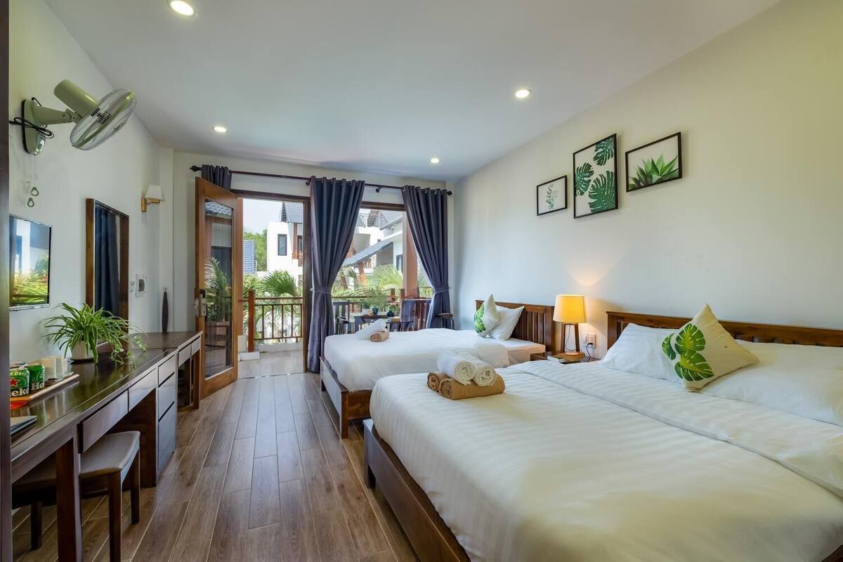 A hotel room with two beds, a desk, a chair, a balcony with curtains, and a view of the outside.