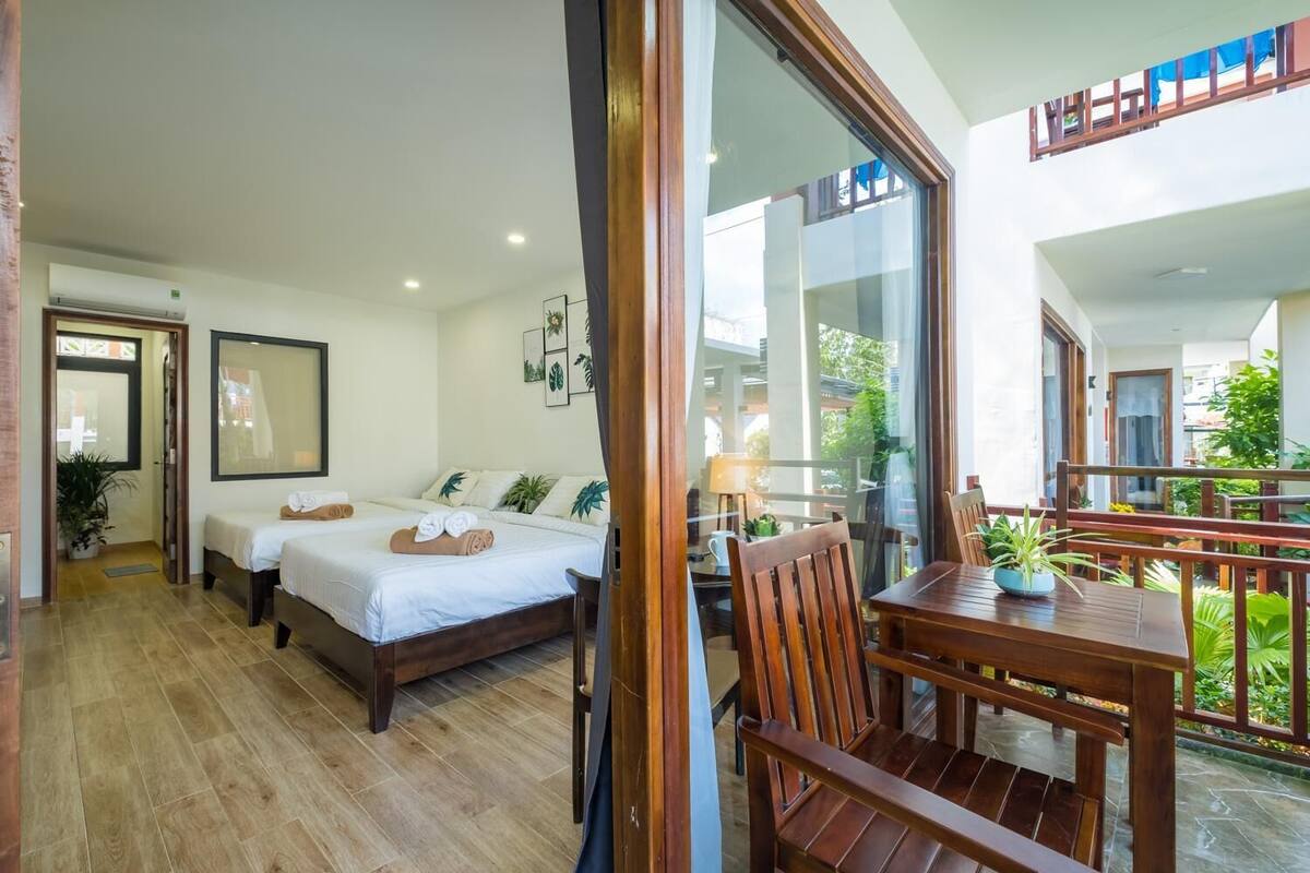 A hotel room with two beds, a balcony with wooden furniture, and a view of the outdoors.