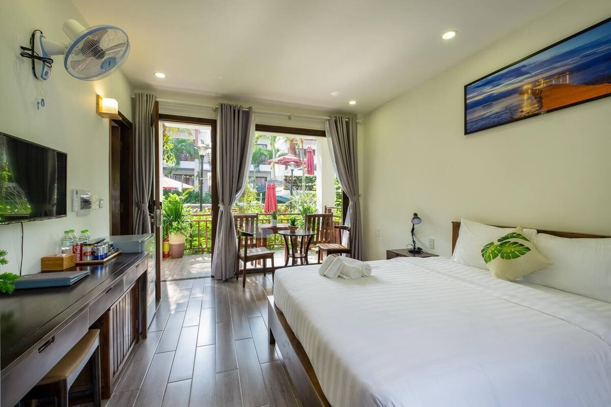 A hotel room with a bed, a desk with a TV, a balcony with a table and chairs, and a painting on the wall.