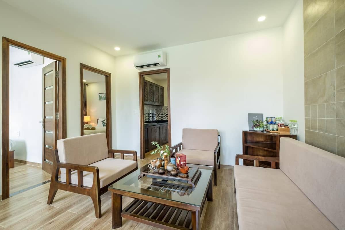 A modern living room with a glass coffee table, two armchairs, a sofa, and a kitchen visible through an open door.