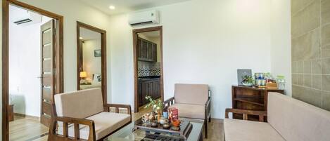 A modern living room with a glass coffee table, two armchairs, a sofa, and a kitchen visible through an open door.