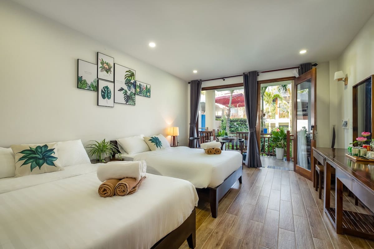 A hotel room with two beds, a TV, a desk, and a balcony with plants.