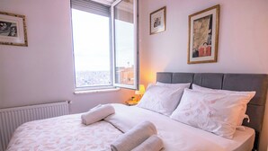 A neatly made bed with white linens and rolled towels, a window with a view of buildings, and two framed pictures on the wall. - Gorgeous Two Bedroom Apartment in Taksim - 3AC (Istanbul)