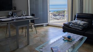 Un salon comprenant une table basse en verre, un canapé, un coin repas avec une table et des chaises, et une grande porte-fenêtre coulissante donnant sur un balcon avec vue sur la mer.