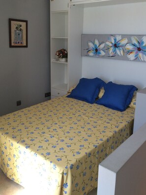 A bedroom with a bed, a nightstand, a shelf with a vase of flowers, and a painting on the wall.