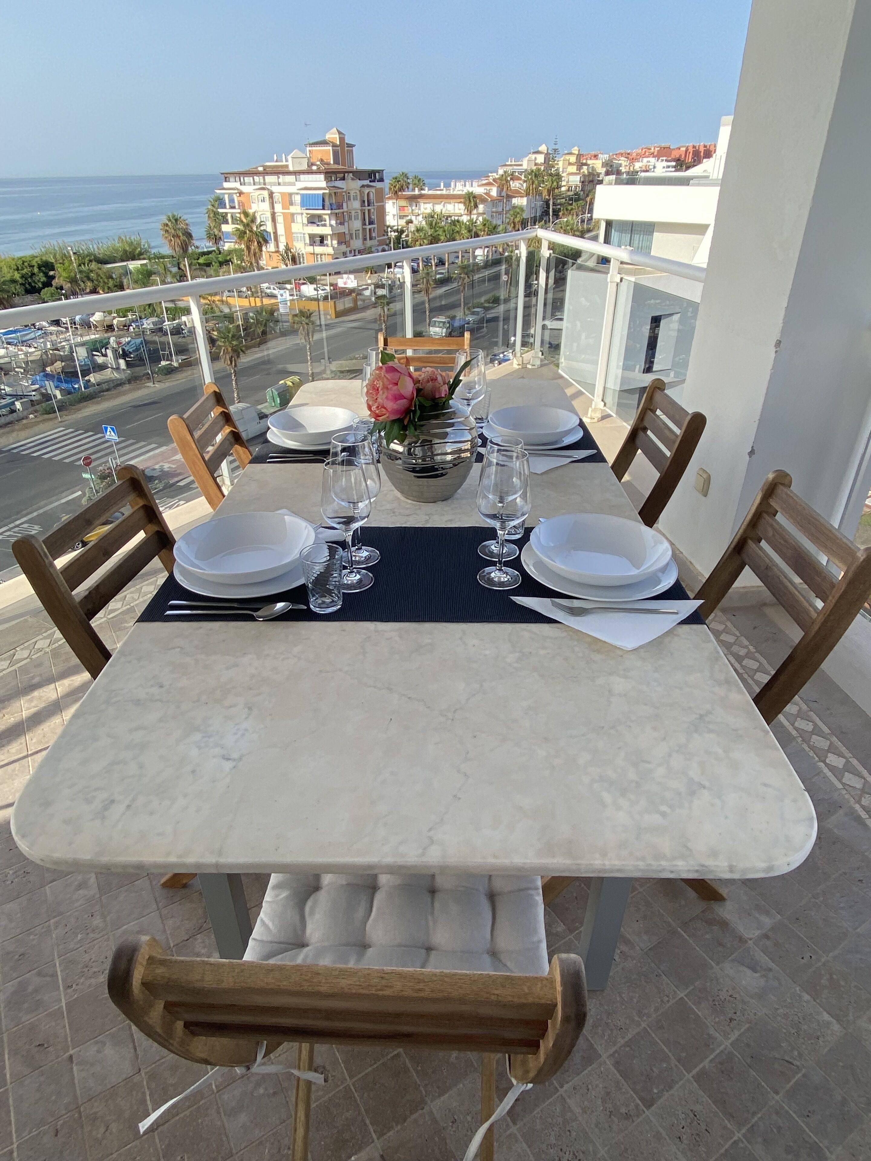 A dining table set for four on a balcony with a view of the sea and buildings.