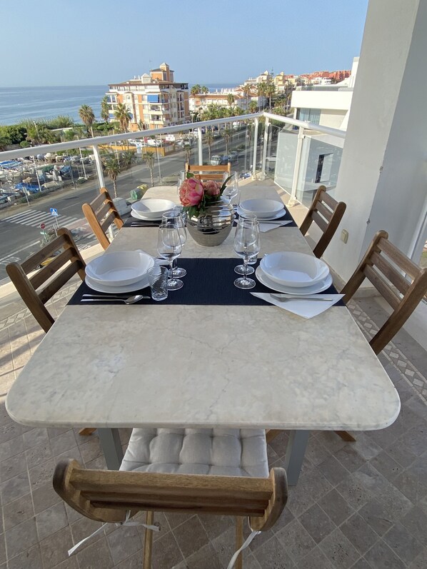 A dining table set for four on a balcony with a view of the sea and buildings.