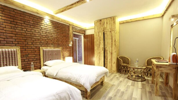 A hotel room with two beds, a brick wall, wooden paneling, a small round table with chairs, and a desk with a lamp. - Silu Homes (Lalitpur)