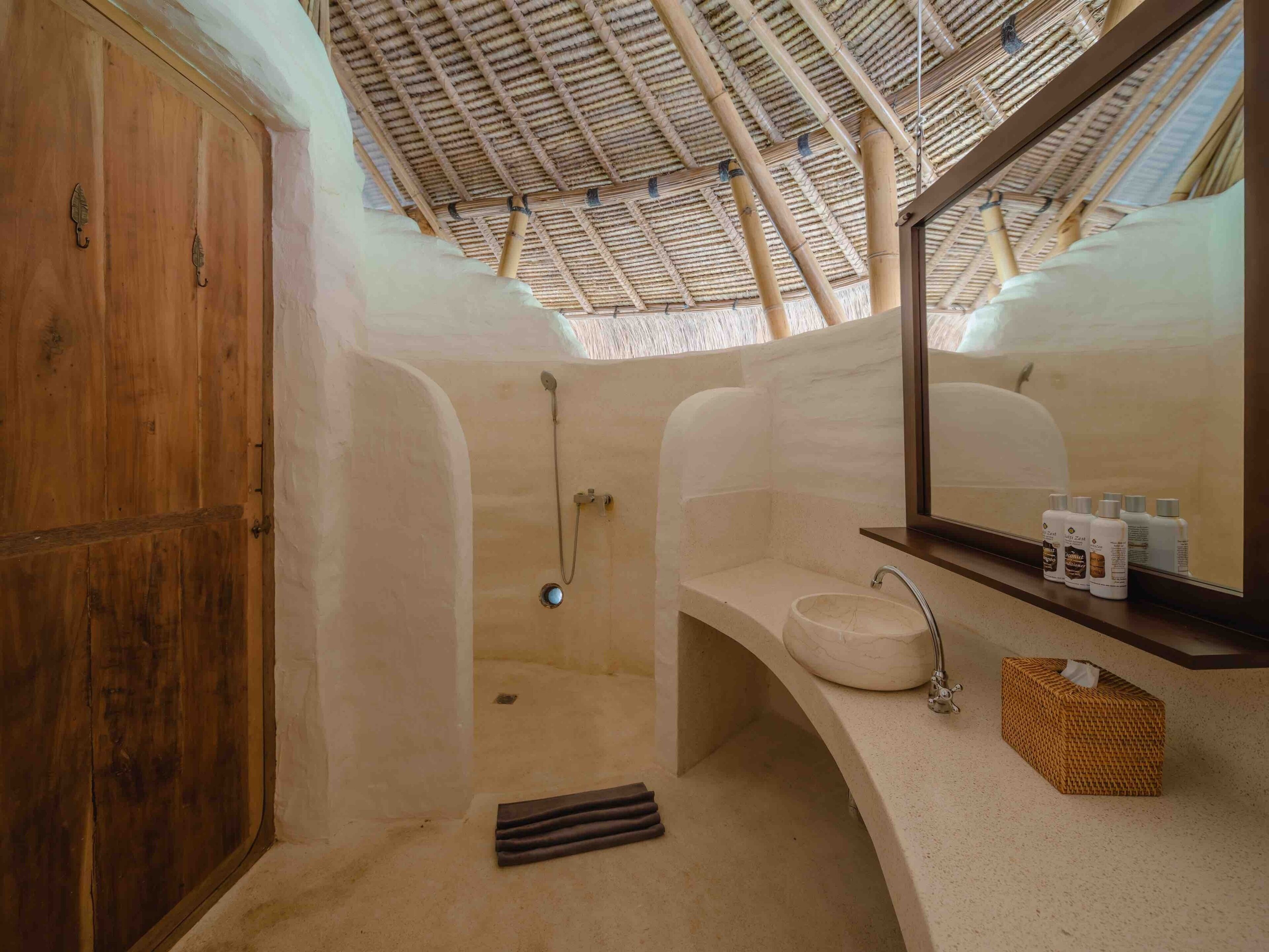 A bathroom with a wooden door, a shower with a bamboo ceiling, a sink with a mirror, and toiletries on the counter.