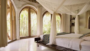 A bedroom with a large bed, a bench, and a canopy. There are windows with curtains and a view of greenery outside. - Mana Earthly Paradise (Ubud)