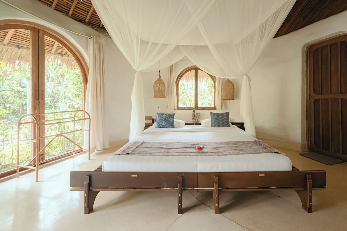 A spacious bedroom with a large bed, a wooden bench, and a balcony with a view of greenery.