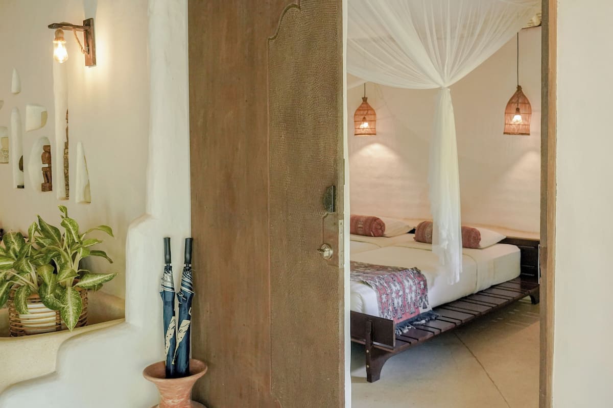 A bedroom with a bed, bedside lamps, a potted plant, and a door leading to another room.