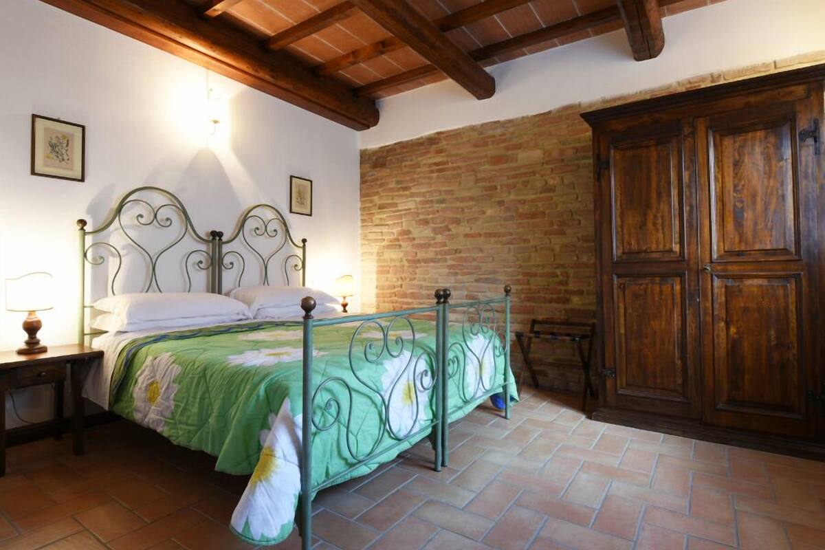 Een slaapkamer met een metalen bedframe, een bedovertrek met groene bloemen, een houten garderobe en een bakstenen muur.