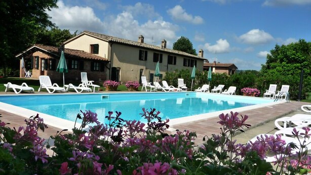 A swimming pool with lounge chairs and umbrellas, surrounded by flowering plants and a building with a tiled roof. - Agriturismo Miralduolo (Torgiano)