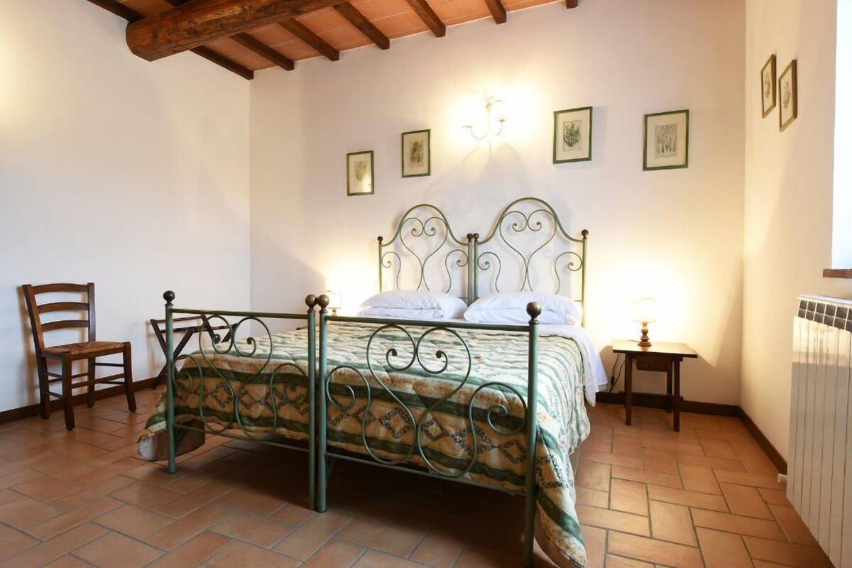 Een slaapkamer met een metalen bedframe, een houten stoel, een nachtkastje met een lamp en een radiator.